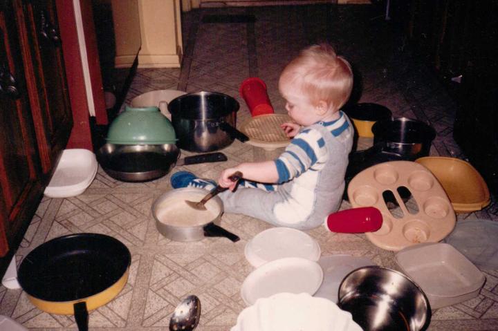 A small child with light pale skin and fair light hair sits on the floor in the kitchen, wearing blue shoes, grey overall sweats, with a white stripe down the side, and a long sleeve shirt with the sleeves rolled up, that is striped blue, grey, and white. they hold a metal spoon in their left hand and are looking down at it as they hit a white metal pot bottom. surrounding them on the floor are the contents of the cabinets and drawers that were pulled out to find the instruments, including a variety of pots and pans, metal spoons, and plastic tupperware, collanders, etc. the floor is a white tan with cross and squared brown tile, with brown wood cabinets off framing the photo.
