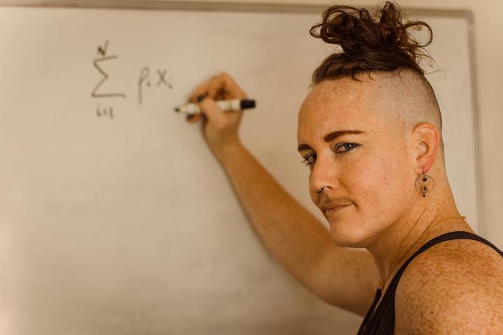 Juniper writing some math at a white board (the sum of rho sub i times x sub i over i from 1 to N) while looking back at the camera over their left shoulder. Their hair is close shaved around the sides and done up in a curly mess. Their eye brows are sharp and they are wearing dangly circle earrings with silver hoops and blue beads. They are giving a smirky smize look at the camera and their freckles are plentiful and multi-colored.