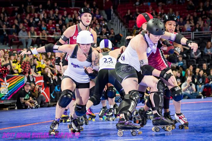 A roller derby game, mid jam, taking place in an indoor arena with fans in the stands and standing behind a short fence with sponsor logos, and photographers on the floor, which is a deep blue with orange track boundaries. Juniper is playing with Rose City, who is in white and purple, competing against Gotham in red and black. Juniper is in the front right of the screen playing offense for Loren Mutch, Rose City's jammer, who is in the center of the photo exploding into a sprint. Behind Juniper directly are three Gotham blockers, and the fourth blocker is behind Mutch, and has a face of slight dejection. As the jammer, Mutch is wearing the white helmet cover with a purple star. Juniper has a black helmet and is wearing a light purple bandana. In the background on the track, you can see Gal of Fray, 23, a Rose City blocker bracing blockers in defense against Gotham's jammer. It is a fleeting moment but a cleanly executed play by Rose City, who would go on to win the semi-final match 159-113. There is a watermark for Adina Butler, IGP Photo 2017 in purple in the lower portion of the picture.