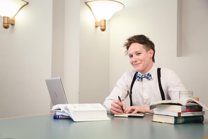 Juniper at a table in a library conference room. She is sitting at the far end, looking across the table at the camera, the table top is pale green with brown trim, the walls are white and plain, with two light fixtures on the walls aiming upwards. Juniper is wearing a long sleeve dress shirt (white with light grey specks), black suspenders, and a blue white and black bow tie. Her hair is relatively short, tapered down to a bizz near her ears, but shaggy and a little mullet-like. She is writing with a black pen in a small notebook and is flanked by propped open text books, a laptop, and a mason jar of water. She is staring at the camera smizing and smirking.